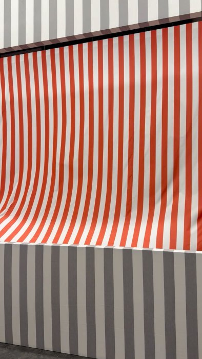 A piece of red-and-white striped cloth (blown by an unseen fan) is cut out from a wall of gray and white stripes.