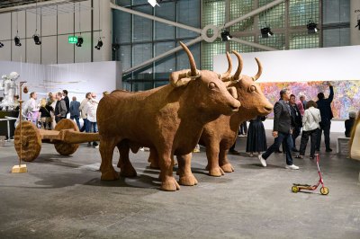 A small red scooter stands in front of a wooden sculpture of two oxen pulling two wheels.