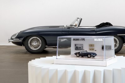 A replica of a navy sports car sits on a pedestal in front of the car that inspired it.