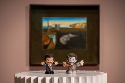 Two Salvador Dalí figurines sit on a pedestal in front of a Dalí painting.