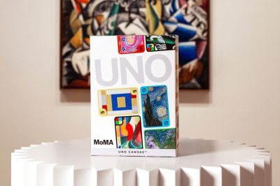 A box for an Uno deck sits on a pedestal in front of a painting that is printed of several of the cards.