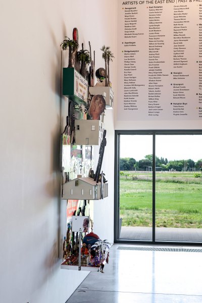 A view of the side of a wall-hung work that is a sculpture with buildings and people popping out.