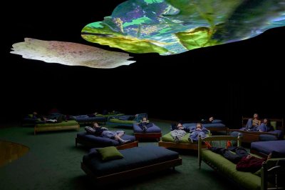 A group of people lying on beds beneath projected abstractions on blobby screens hanging from a ceiling.