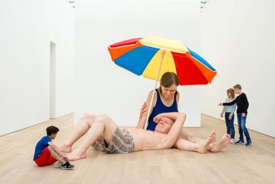 A large-scale sculpture of a reclining man and a seated woman in a bathing suit beneath a beach umbrella. Smaller people point to the work.