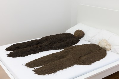 A bed with two dirt forms resembling human bodies on it. The forms have stones for heads.