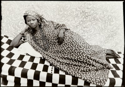 A photograph of a Black woman in a floral-print dress reclining on a checkerboard blanket.