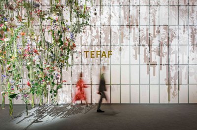 Two blurred figures walk past a wall that says TEFAF. At left is a suspended installation containing various flowers and other plants. 