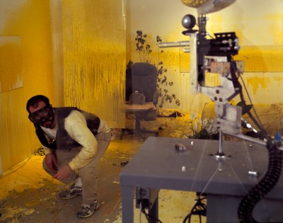 A brown man with ski goggles on dodges a paintball gun in a room covered in yellow paint.