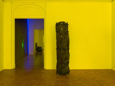 A sculpture resembling an object contained in knotted black latex in a room lit in yellow.