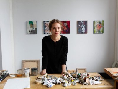 A woman standing at a table with tubes of paint on it. She stands in front of five abstract paintings.
