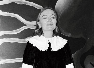 A black-and-white photograph of a white woman smiling in front of an abstract painting.