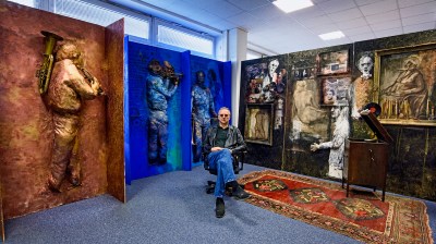A photograph shows the artist Predrag Đaković in his studio, seated, in jeans, leather jacket, and sunglasses. Behind him are a number of artworks, mostly paintings with some three-dimensional elements including human figures, some playing musical instruments.