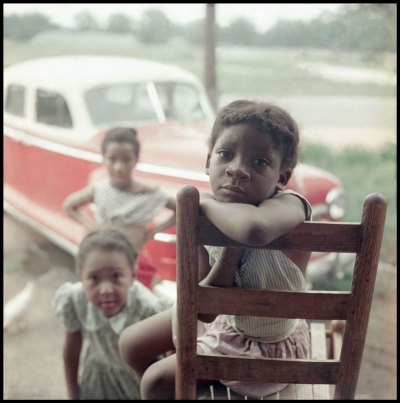 Three Black children stare out at you. One is seated in a wood chair and is the only one in full focus. Behind them is a red muscle car. 