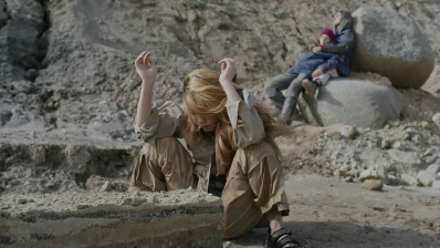 A film still showing a woman crouched down with her ams up in a desert landscape with two people behind her leaning on rocks.
