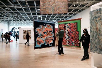 People walking amid freestanding abstract paintings suspended from a grid of metal ceiling beams.