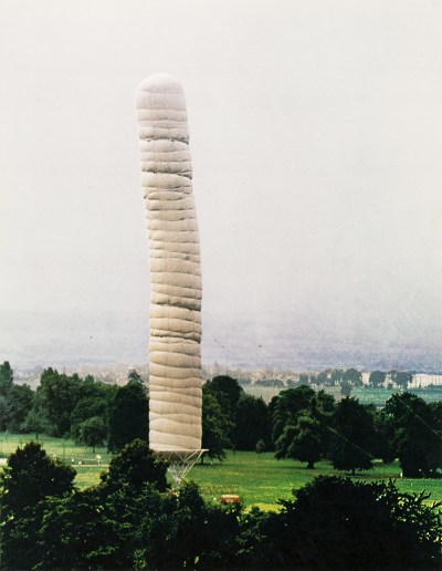 a tall white inflated column in a green field, towering above the trees with a gray sky behind
