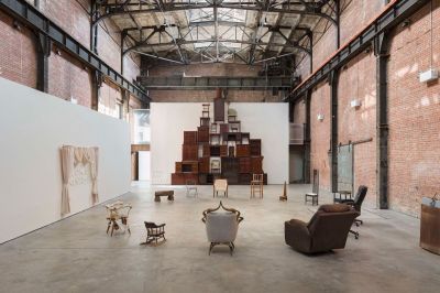 A ring of stools and chairs arrayed in a U shape in the center of a gallery. Behind the installation is another composed of stacked cupboards and cabinets.